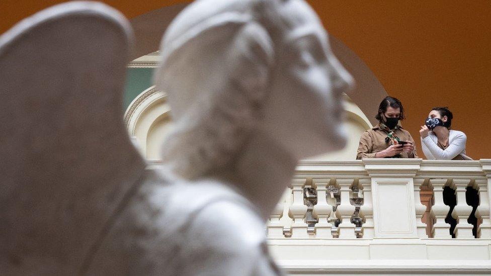 Two women wear masks as they chat with a marble statue in the foreground