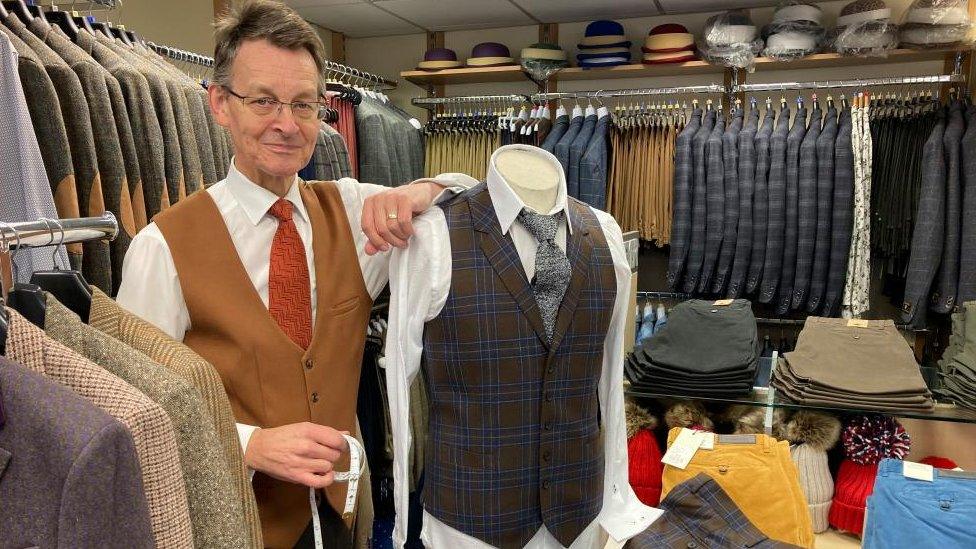 Man wearing brown waistcoat and red tie stands next to a mannequin wearing a grey waistcoat and tie