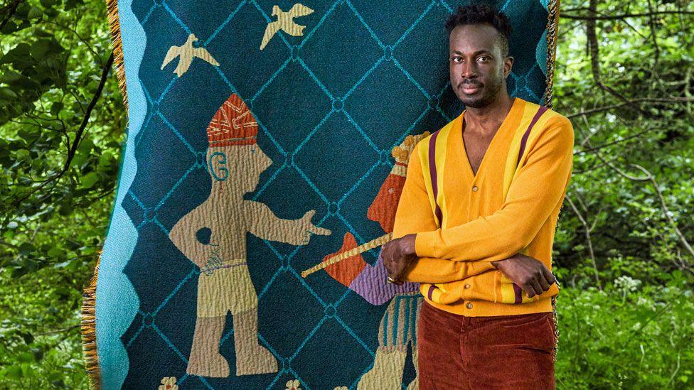 Cameroonian-Nigerian interior designer Amechi Mandi, dressed in a yellow cardigan and burnt brown cords, stands in a garden with arms folded in front of one of his throw blankets called 'Chief Palaver' showing two figures and two birds stitched on to blue diamond designed thick woven fabric 