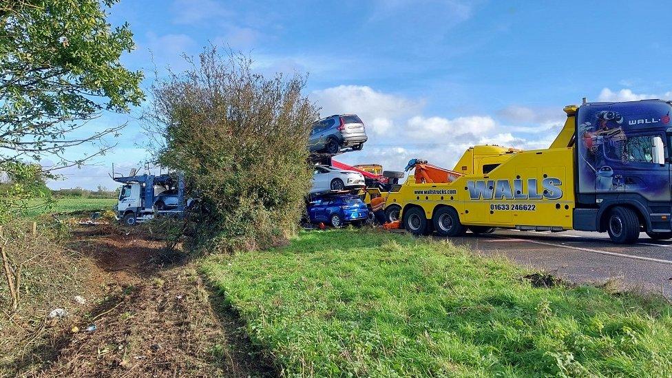 Car transporter stuck in some mud in a field, with a large recovery vehicle behind it dragging it out.