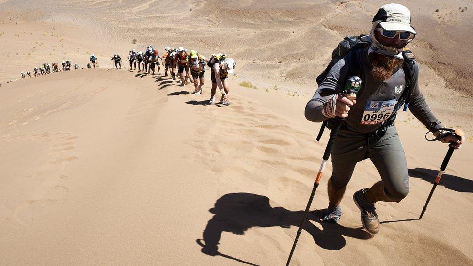 A-man-treks-ahead-of-a-line-of-people-up-a-massive-sand-dune-in-the-Sahara-Desert.