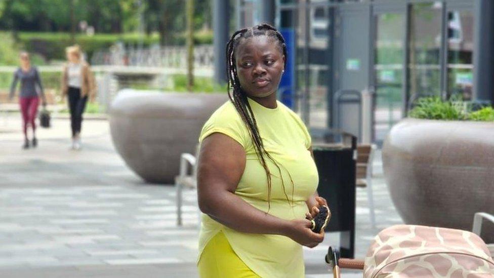 Mary Kukua Anderson stood next to a child's pram. She is wearing all yellow and has her hair in braids.