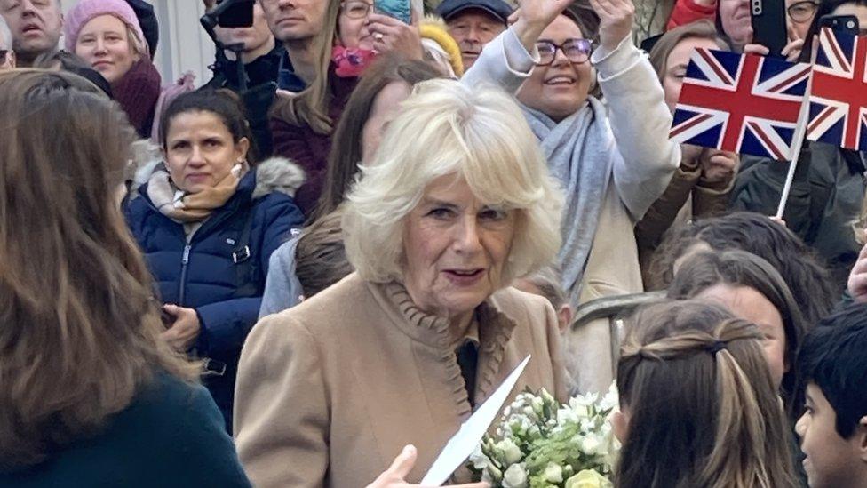 The Queen holds a bouquet of flowers, surrounded by people, with a couple waving union jack flags