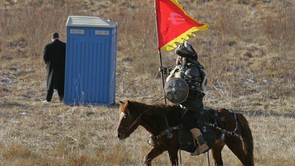 An actor dressed as a Mongolian warrior rides his horse past a US Secret Service Agent standing next to a portable toilet