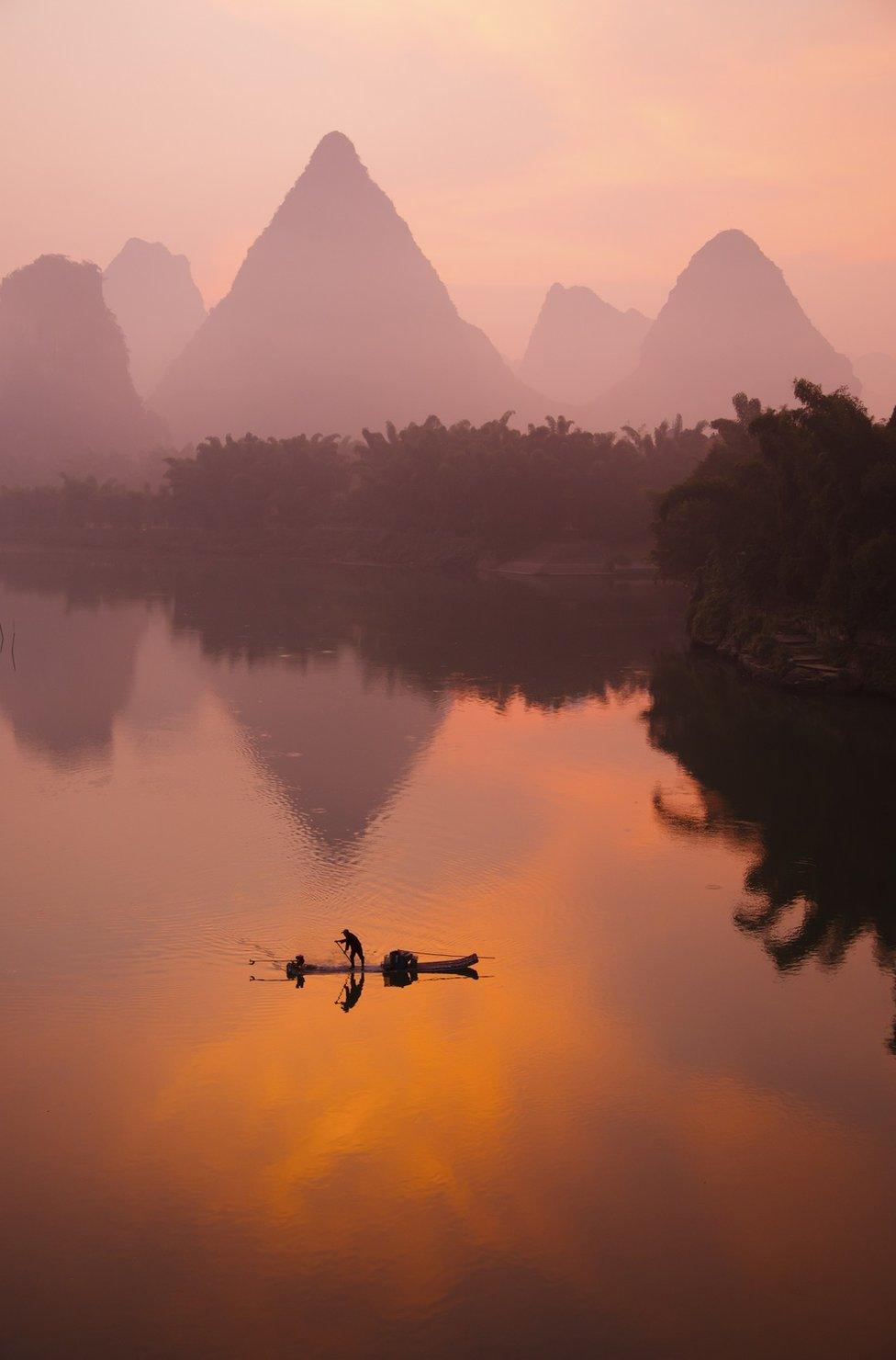 A sunset view of a person on a boat on water with mountains behind them