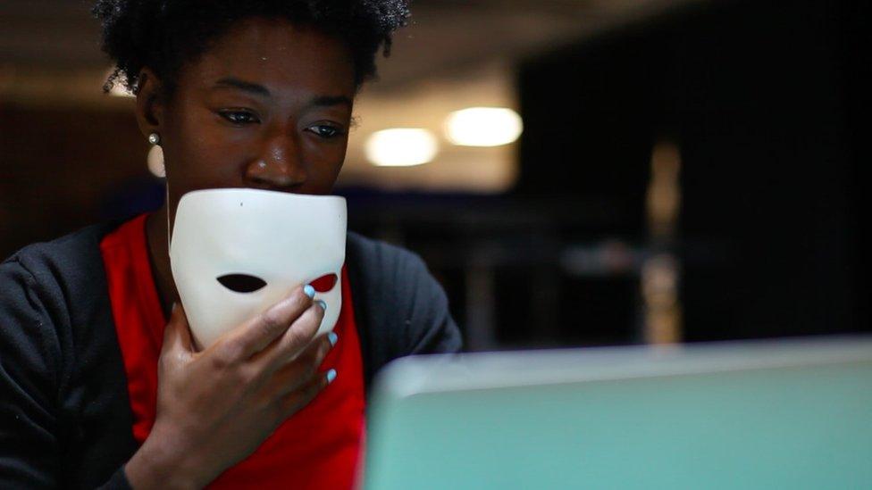 Joy Buolamwini holding a white mask at a computer