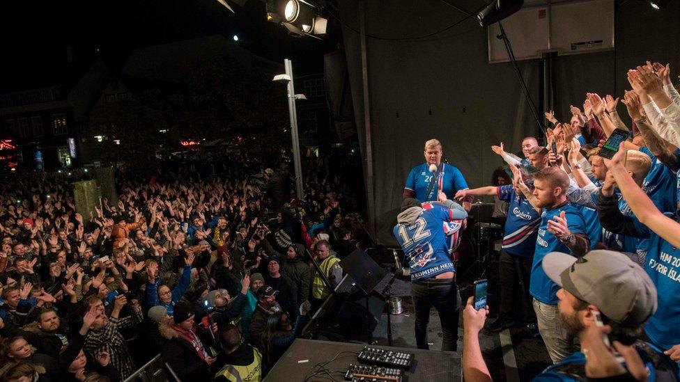 The Iceland team dressed in blue hold their arms open wide on a stage as fans cheer below.