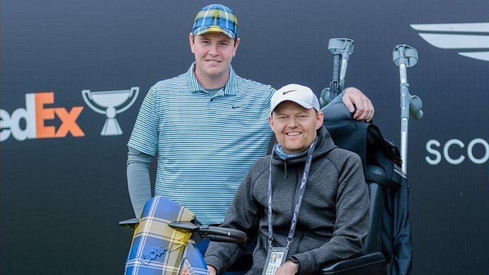 Robert MacIntyre is standing beside Scott who is sitting in a wheelchair. Robert is wearing a blue and yellow checkered cap and stripy blue top. Scott is wearing a white Nike cap and grey hoodie.