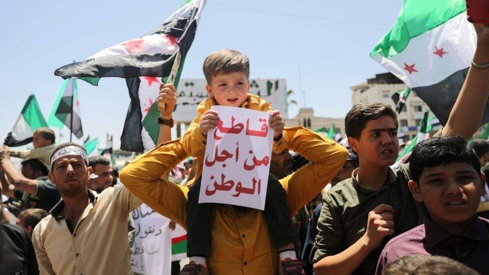 A boy, carried on the shoulders of a man, holds a sign reading "boycott for the sake of home" during a protest against the presidential election in opposition-held Idlib province (26 May 2021)