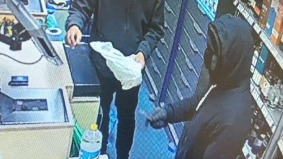 Gawthrop in a black hoody holding a knife towards a male member of staff behind the till.