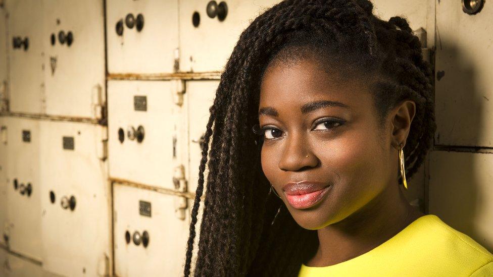 A young black woman with hair styled in long locs parted at the side leans against a bank of cream-coloured drawers that look like a filing system of some sort. She's smiling, wearing a bright yellow top and dangly gold earrings.