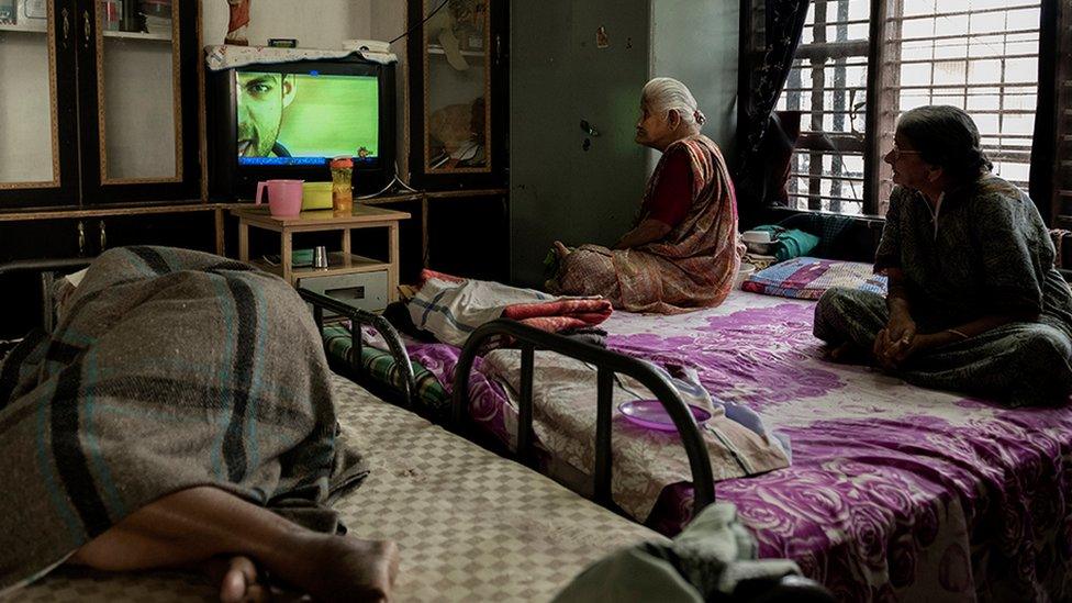 Two old women watching TV while an older person sleeps on a bed near them