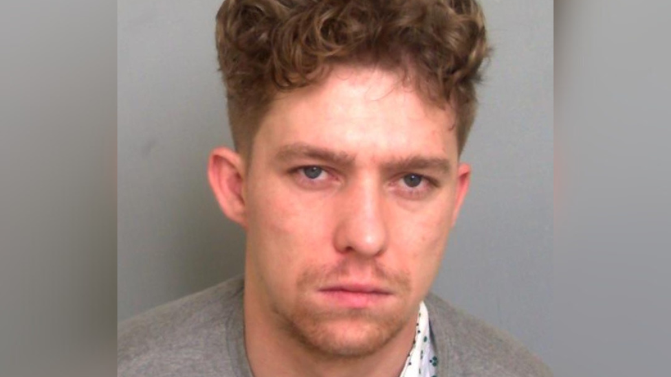 Custody mugshot of Michael Burke. He has curly brown hair. He is wearing a grey sweater and has a white bandage on his neck.