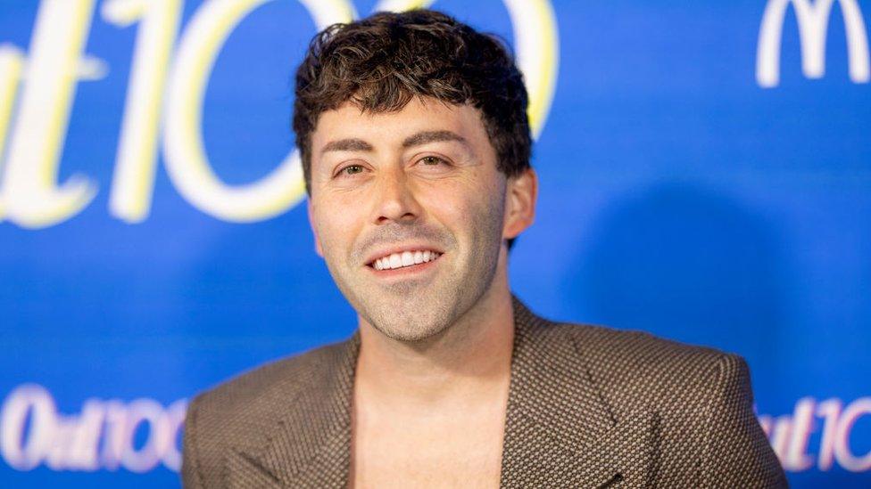 Singer-songwriter Leland on the red carpet for the OUT100 Party in Hollywood. He is stood in front of a bright blue background and is smiling for the camera. He has short dark hair and stubble and is wearing a brown blazer that is open showing his chest.