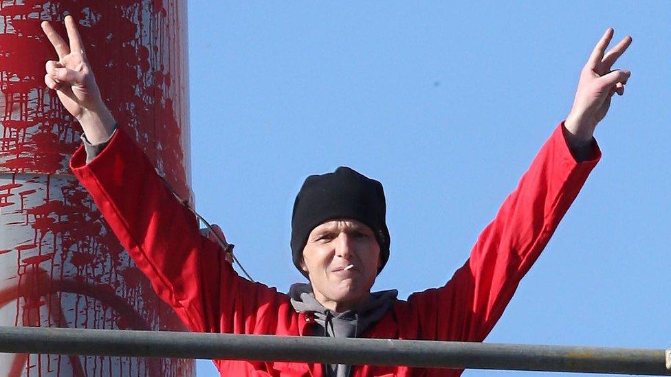 A man in red overalls and a black hat with a cigarette in his mouth hold his hands aloft and gives peace signs with his fingers