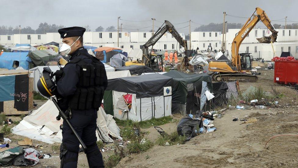 A police officer stands guard while mechanical diggers start to crush shelters in the makeshift migrant camp known as the Jungle near Calais, northern France, on 26 October 2016