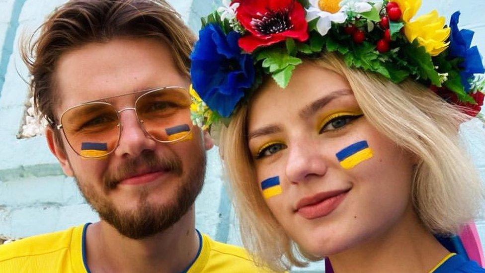 Serge and Polina, a Ukrainian man and woman. Serge on the left is wearing sunglasses with blue and yellow of Ukraine's flag painted under each eye. He is wearing a yellow Ukraine shirt. Polina also has the blue and yellow Ukraine flag paint under her eyes, and has a flowery crown on her head.