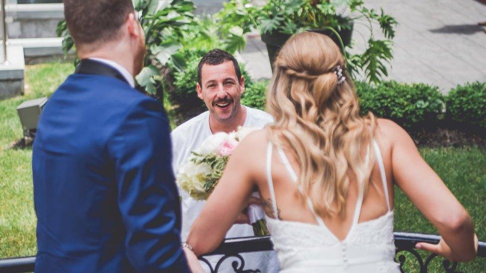 The couple in their wedding attire look over a balcony to talk to Adam Sandler.