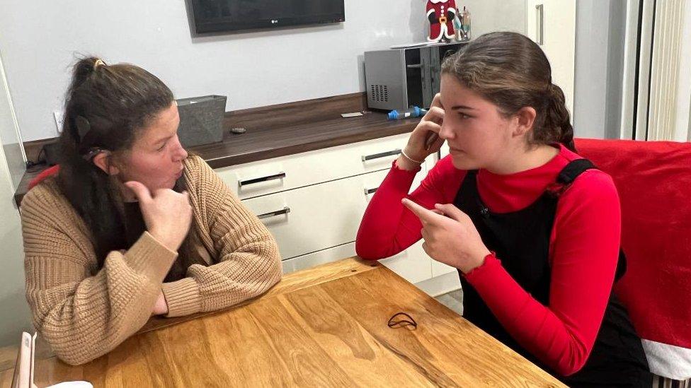 Woman in brown top sits at a table with a girl holding a phone