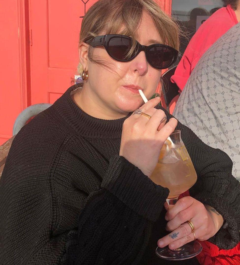 Martha Smith is wearing a black jumper and black sunglasses and is drinking from a glass and sitting in front of a red door.