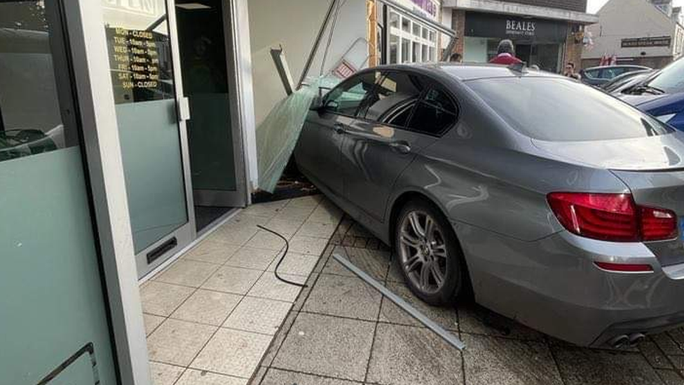 A grey BMW partially inside a tattoo studio, a knocked out window frame with shattered glass and tyre tracks on the pavement.