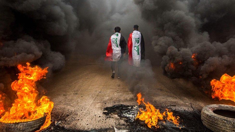 Anti-government protesters wearing Iraqi flags walk past burning tyres in Basra, Iraq (17 November 2019)