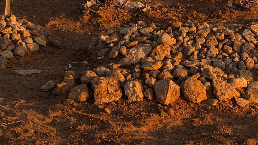 Graves covered by cairn stones litter the ground outside a village or camp seen in the distance