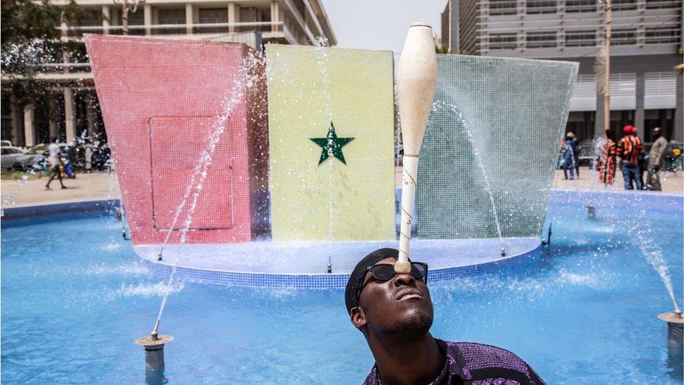 Man balancing a sports' bat on his nose. He is outside. Behind him is a Senegal flag.