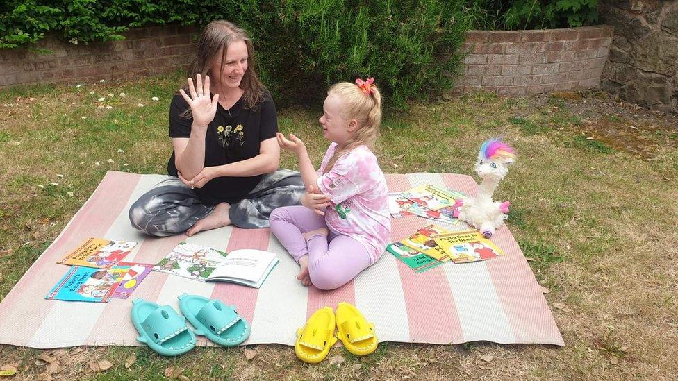 Nikki and Lucinda sit on a picnic blanket and sign to each other.