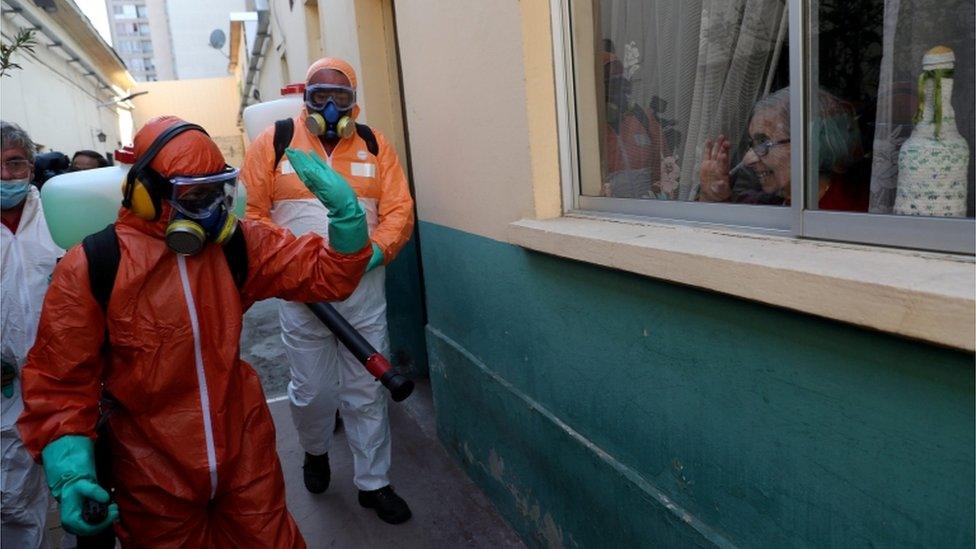 A worker wearing protective gear waves to a woman while using disinfectant to clean outside of a house in Santiago, Chile