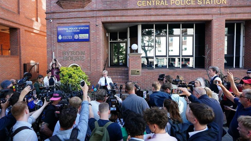 A police officer addressing a large crowd of journalists outside a police station.