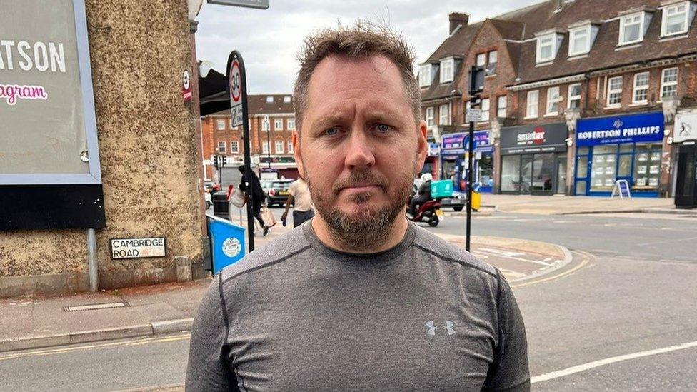 Alex Mitchell, a man with brown hair wearing a grey sports top, standing by the advert in Harrow