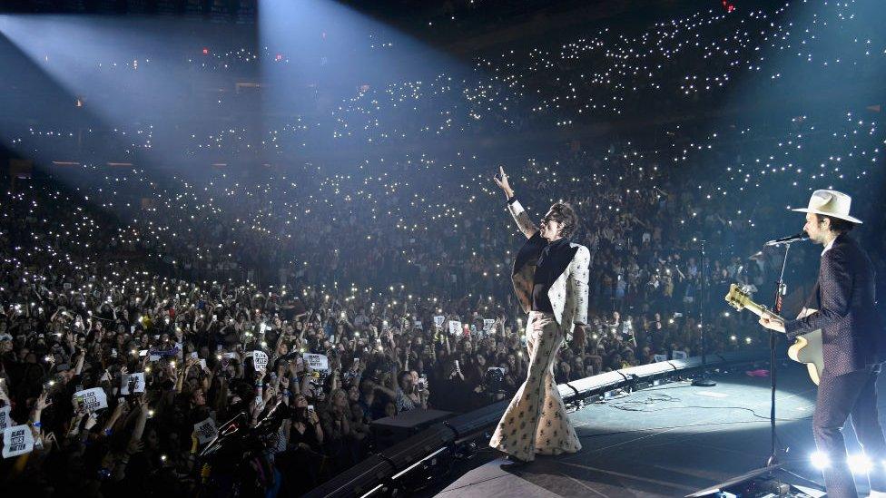 Harry Styles in front of a large crowd, holding phones