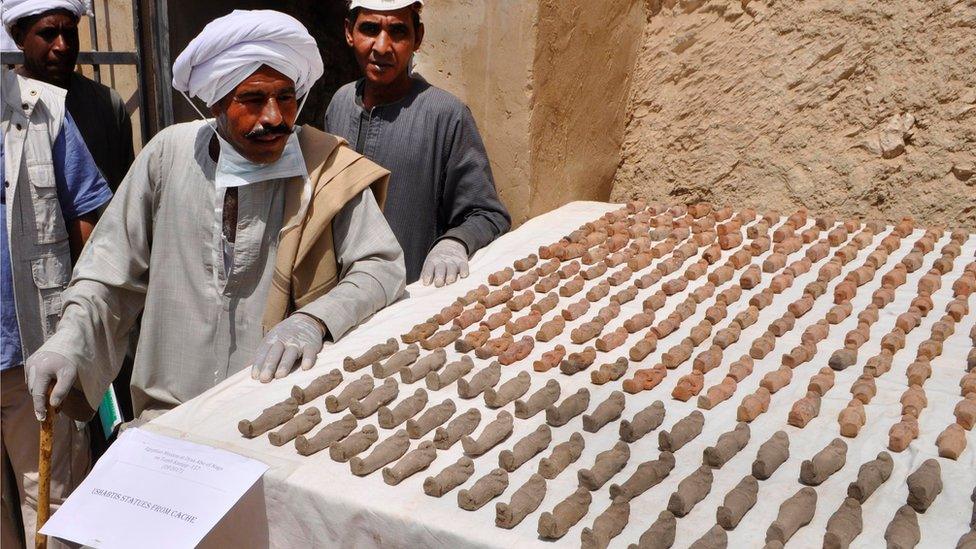 Two men next to a table covered in rows of clay humanoid figurines