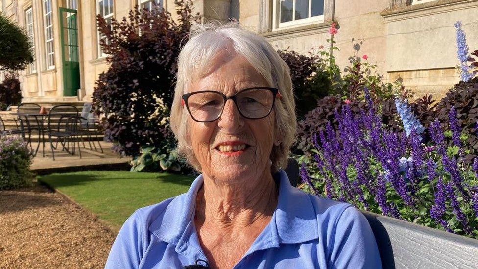 Woman with light-coloured hair wearing blue shirt sites on a bench with stately home behind her.