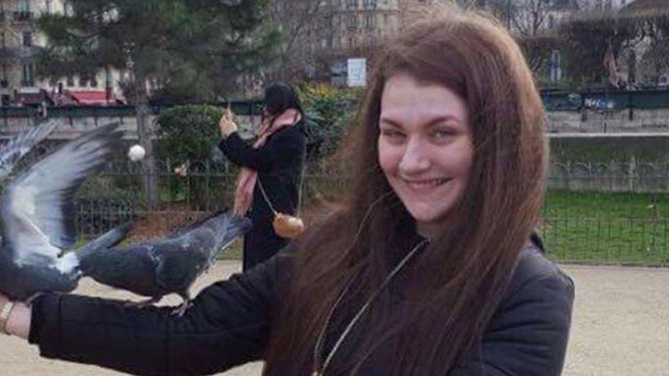 Libby, who has long brown hair and wearing a black coat, with two pigeons on her right arm standing in a gravel and lawned park area