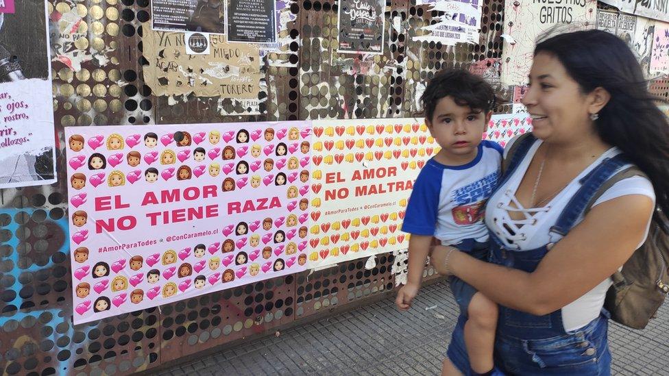 A woman with a child walks past a wall with posters saying "love does not have a race"