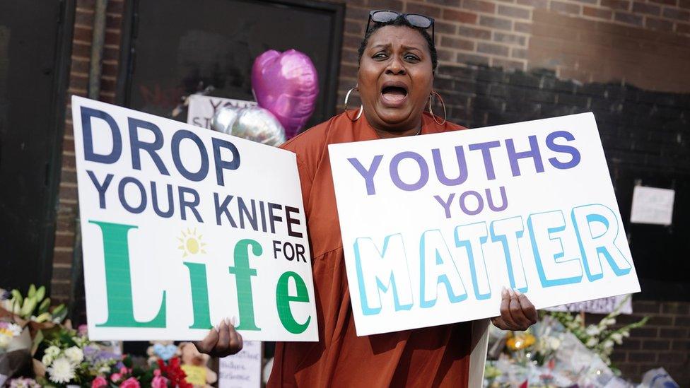 A woman holds signs in front of flowers left in memory of Elianne Andam