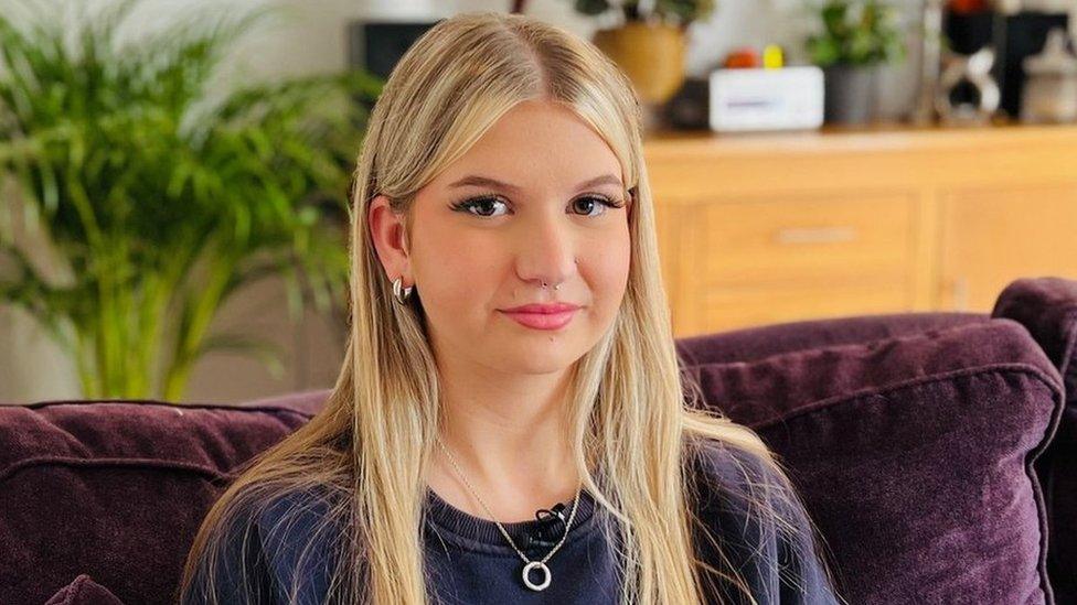 Keira sitting on a purple sofa with a big green plant in the background and a brown wooden unit. She has blonde hair and has her nose and ears pierced. She has a ring on a necklace around her neck and has a purple jumper on.