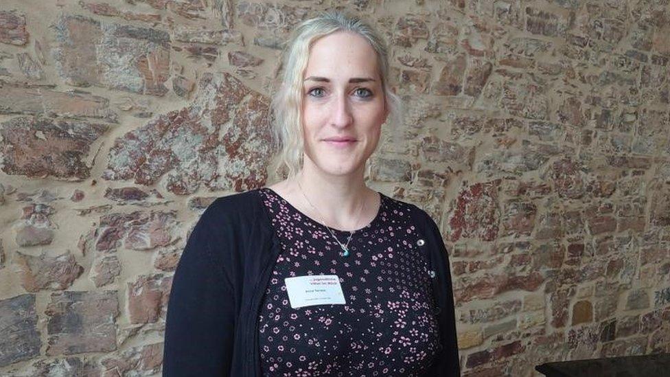 A photo of professor Anna Tarrant stood in front of an old-fashioned looking stone wall. She has light blonde hair and is wearing a black floral top with a black cardigan. She has a silver necklace on and a white name sticker on her top that we can't make out the wording on.