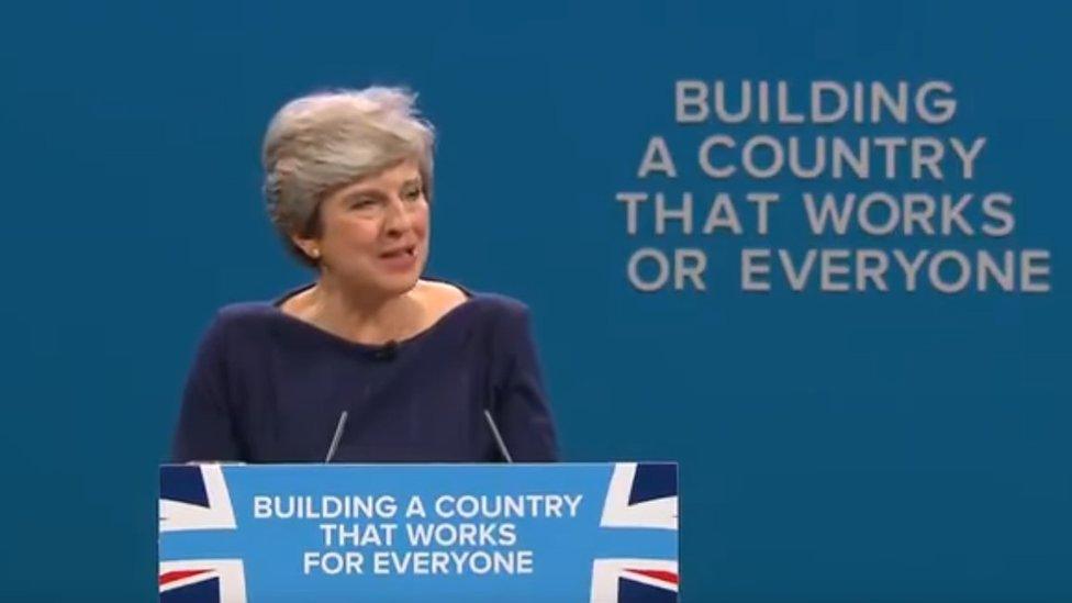 Theresa May speaking as a letter falls off the slogan behind her