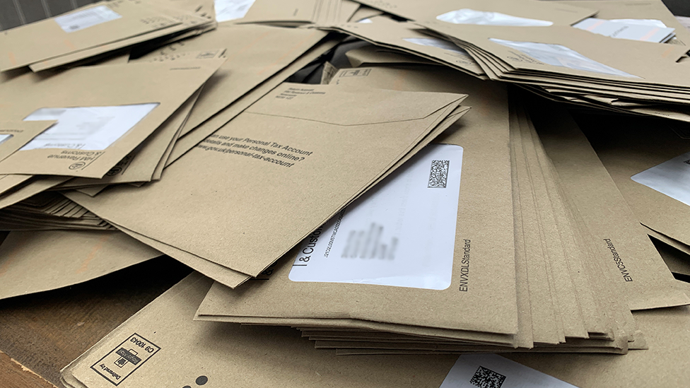 Dozens of brown envelopes with a white address windows piled up on a desk