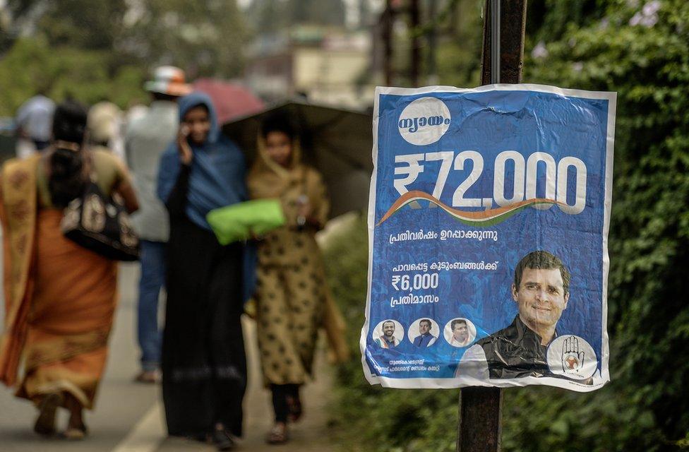 People walk past a poster of the Congress party's minimum income guarantee scheme for the poor