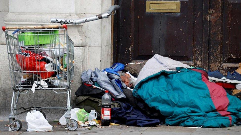 Sleeping bag and other belongings on the floor in front of a wooden door. A shopping trolley of belongings and a cola bottle are visible