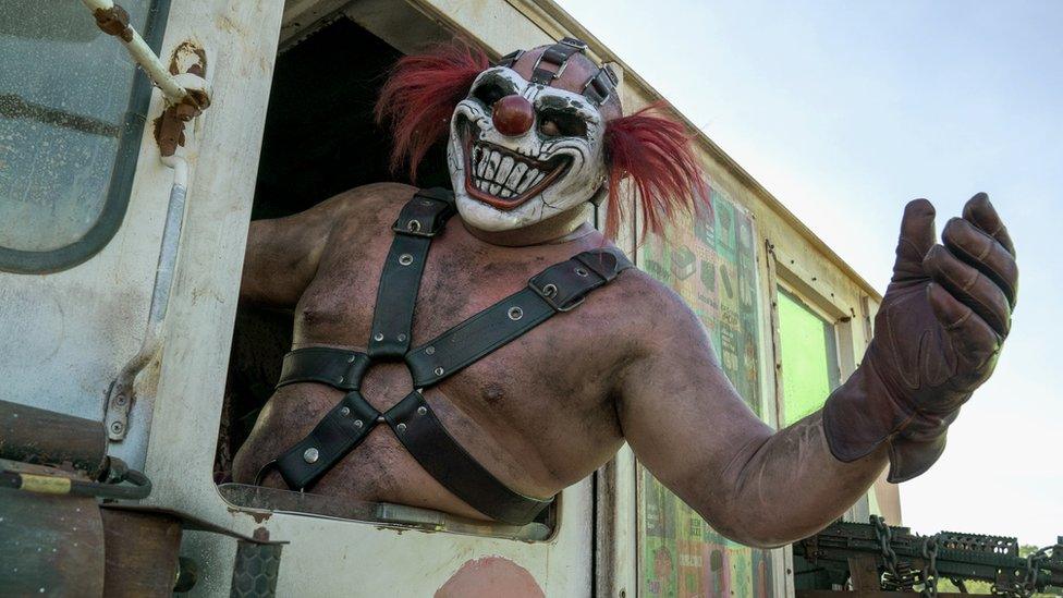 A burly, shirtless man wearing an evil clown mask - with tufts of thick red fake hair sprouting out of each side of the head - leans out of the window of a very dirty ice cream truck. He's shirtless but wears a leather harness with five straps attached to a circular anchor piece over his sternum. He wears a large leather glove on his visible hand. Like the truck, he also appears to be covered in a film of dirt or grease. The mask's exaggerated, toothy smile lends the picture an air of menace