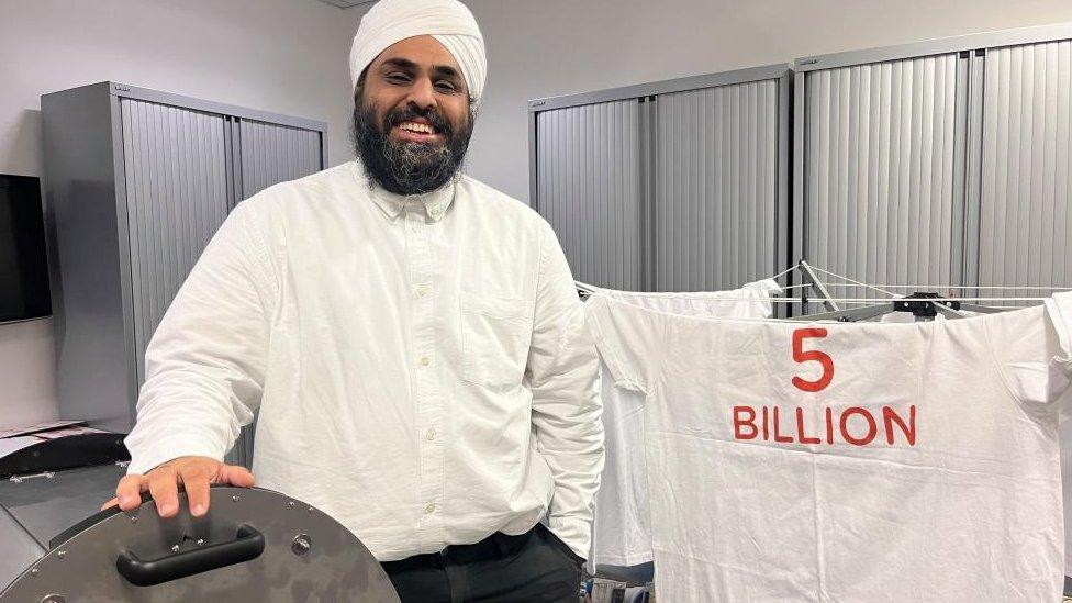 Man with beard wearing turban and standing next to washing machine and t-shirt which states "5 billion"