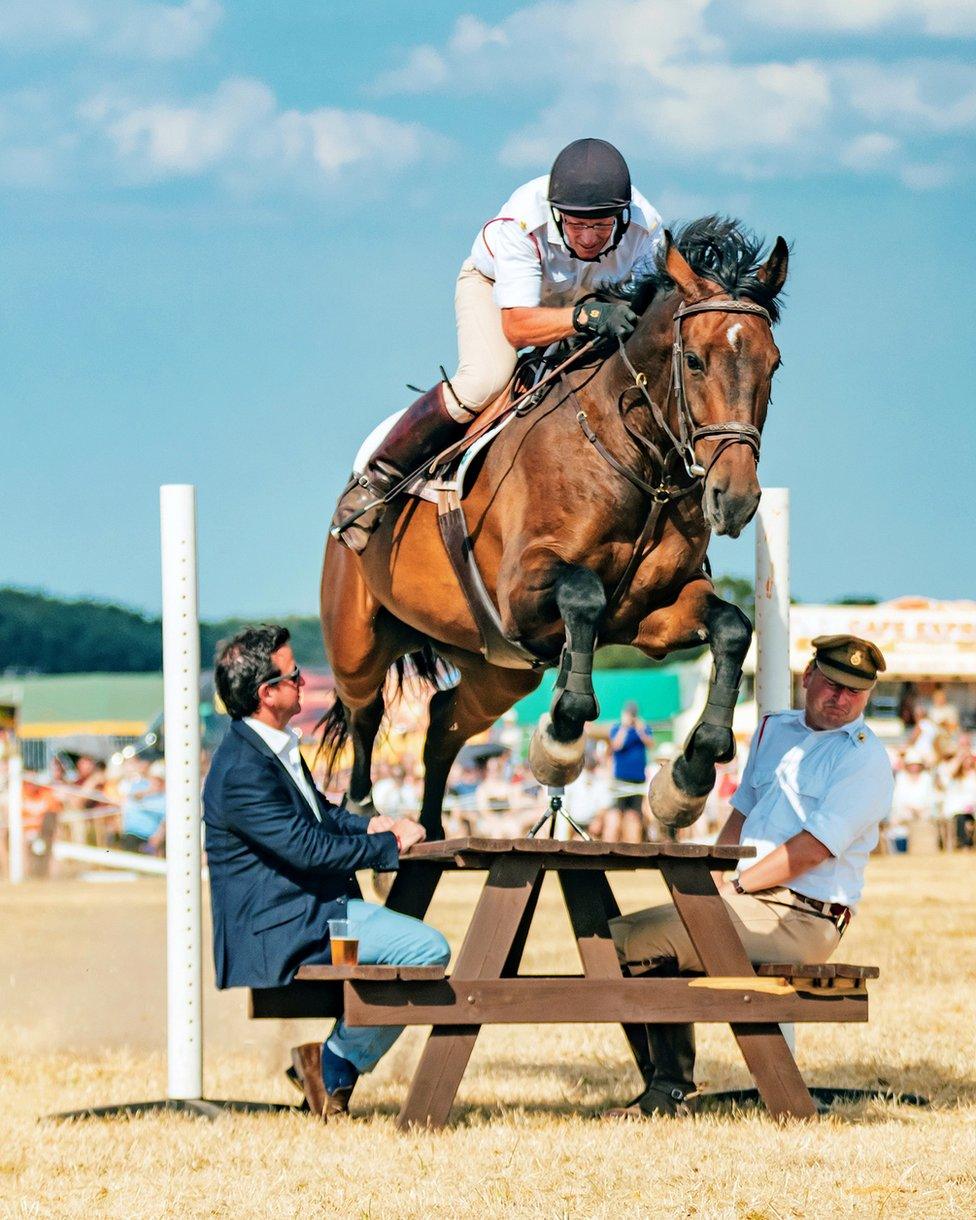 A person on horseback leaping between two men sat at a picnic table