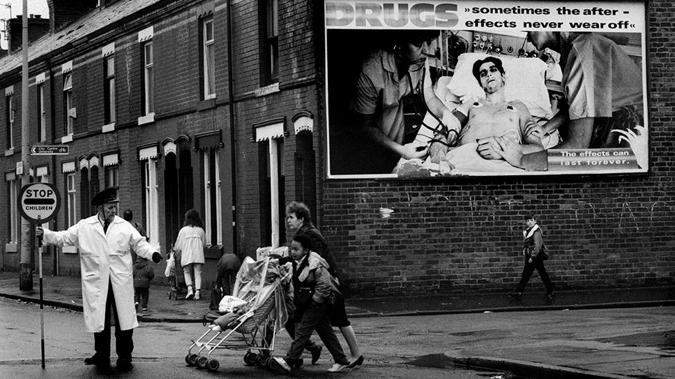 A lollipop man guides children to school across a road in Moss Side