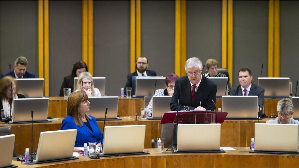 View of a group of Labour Senedd members, including Mark Drakeford, who is stood up making a speech.