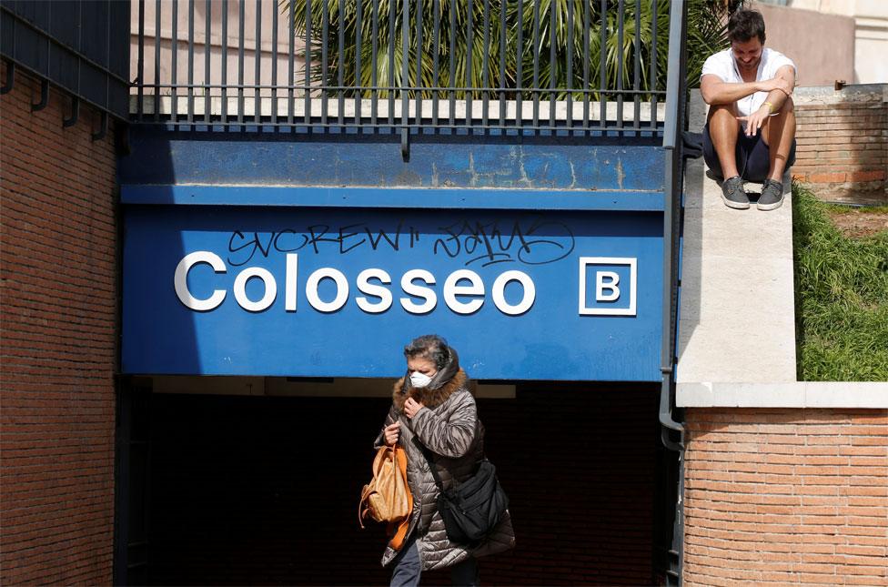 A woman wearing a face mask walks past a metro station whilst a man sits on a wall behind her